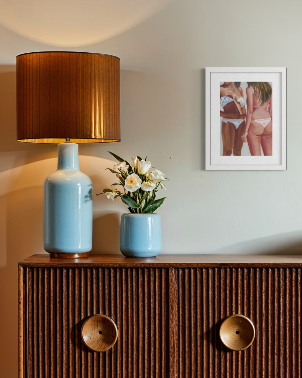A framed art print depicting two women in swimwear hangs on a beige wall above a wooden cabinet. On the cabinet, a light blue ceramic table lamp with a brown pleated shade and a matching light blue vase with white flowers are arranged.