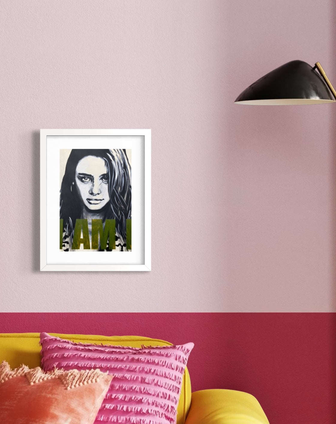 A framed art print featuring a black and white portrait of a woman with green 'I AM' text, displayed on a two-toned pink and red wall above a yellow sofa with pink pillows.