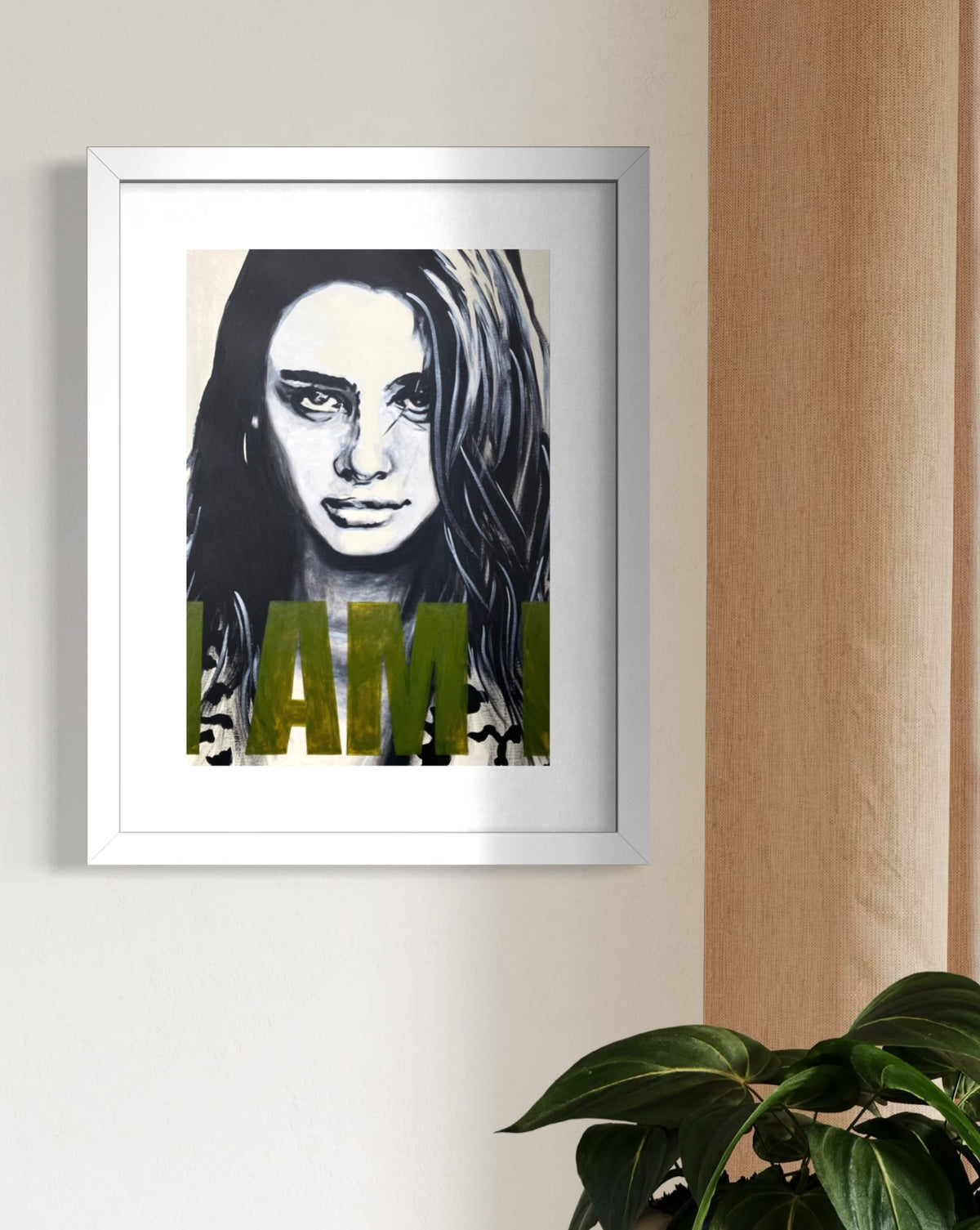 A framed art print featuring a black and white portrait of a woman with olive green text reading 'I AM' across the bottom, displayed on a light beige wall next to a wooden panel and above a green plant.
