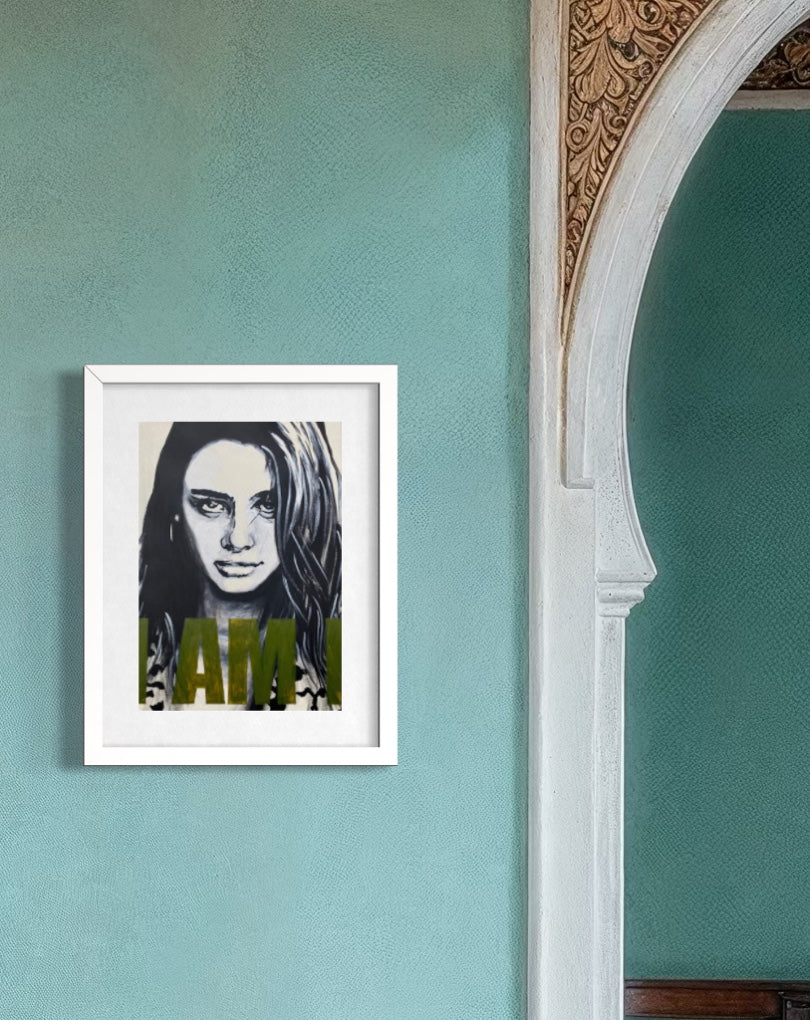 A framed art print featuring a black and white portrait of a woman with olive green text reading 'I AM' hanging on a textured teal wall next to an arched doorway.