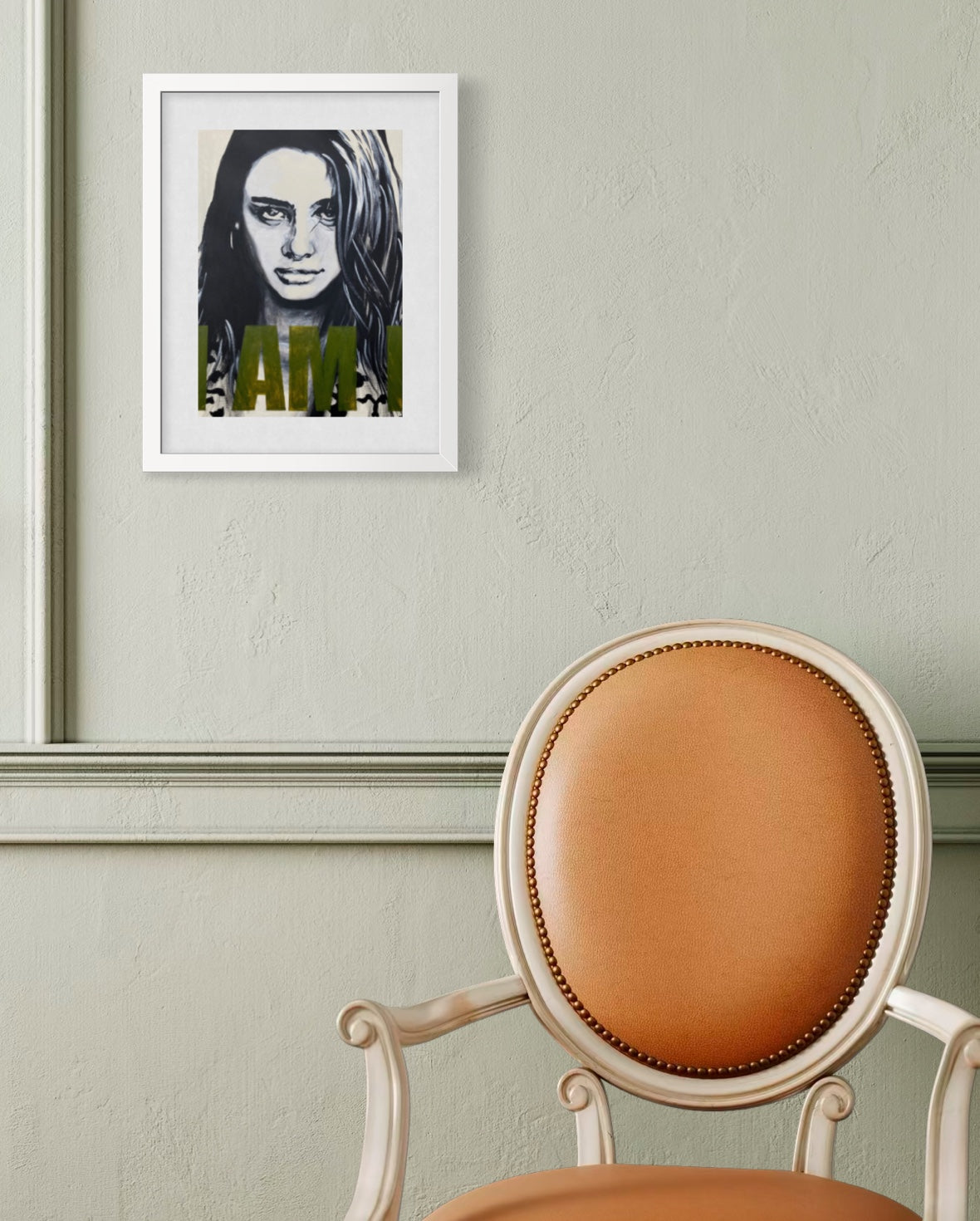 A framed black and white portrait print with olive green text hanging on a textured light green wall above a tan upholstered chair.