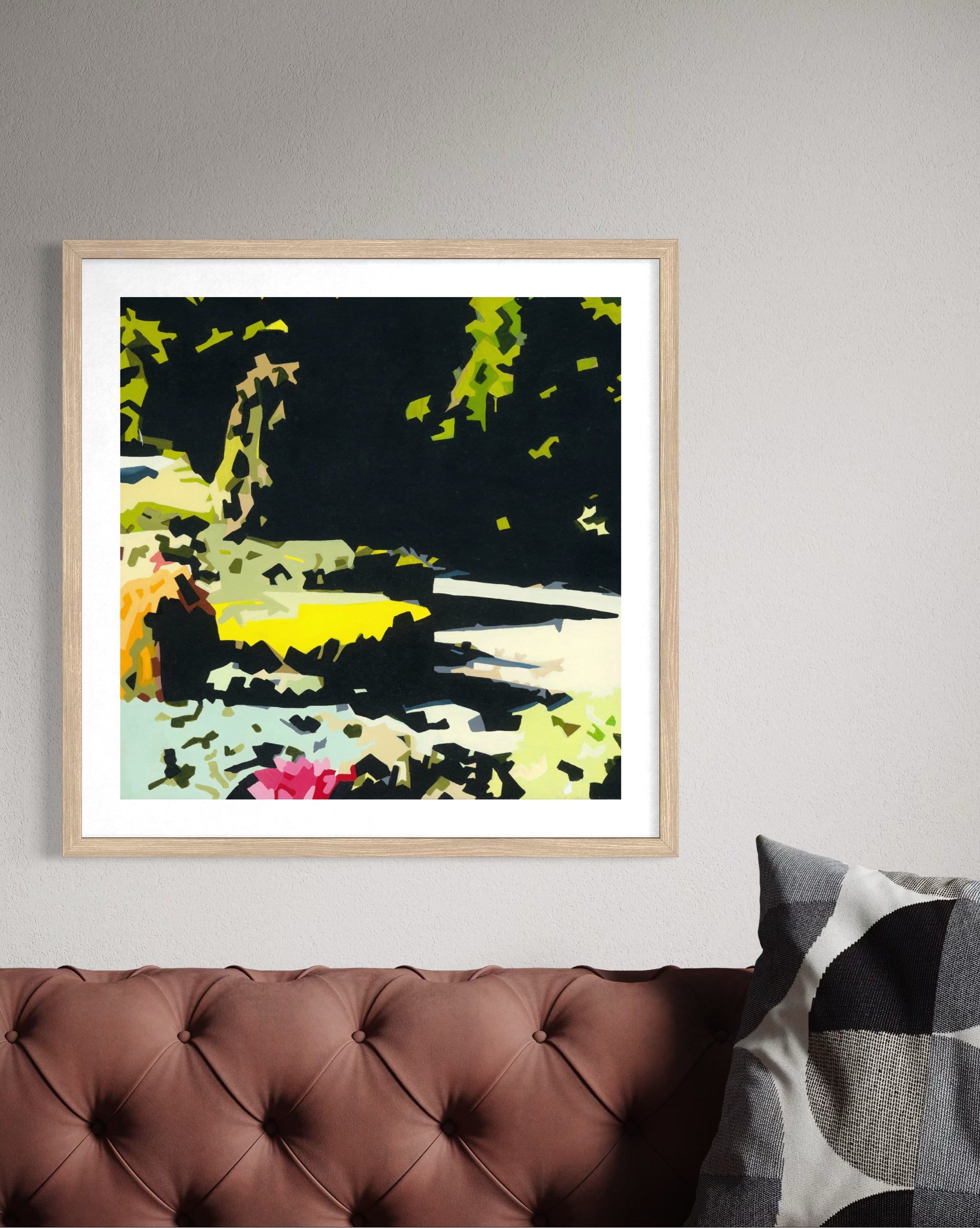 A framed abstract landscape art print with bold black, green, yellow, and pink brushstrokes, displayed on a light gray wall above a brown tufted leather sofa with a patterned pillow.