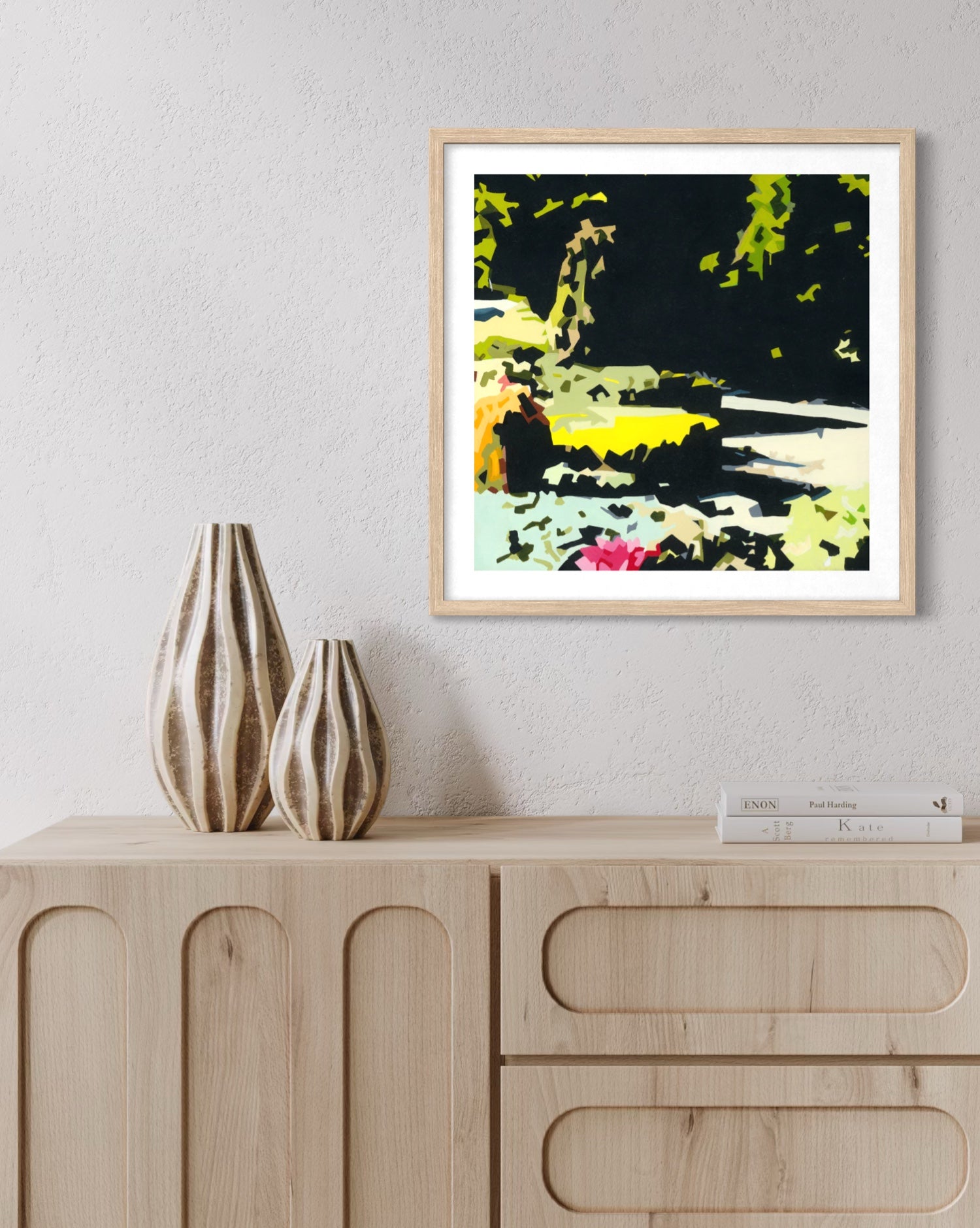 A framed abstract landscape art print with bold black, green, yellow, and pink brushstrokes, displayed on a light gray wall above a light wood sideboard with two ribbed vases and a book.