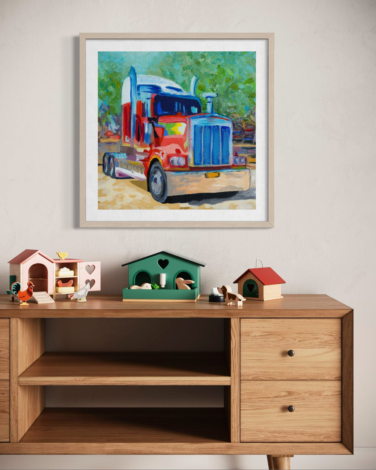 A framed colorful painting of a semi-truck hanging on a light beige wall above a wooden credenza with small toy houses and a green birdhouse.
